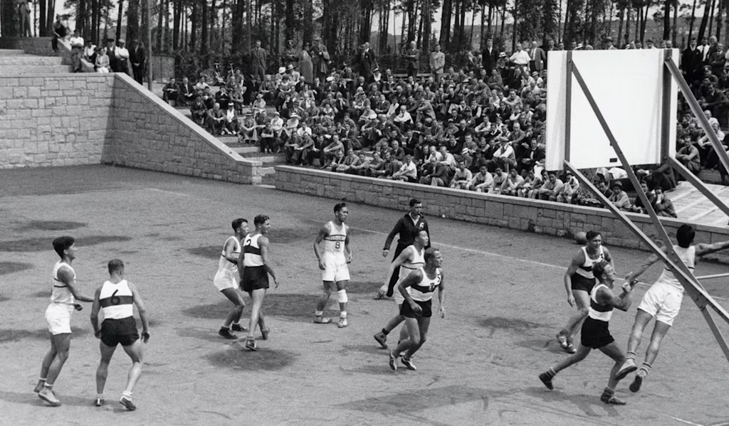 1936년 베를린 올림픽 야외 농구 코트. 모래 바닥에 hoop이 설치되고 관중이 돌계단에 앉아 있다.